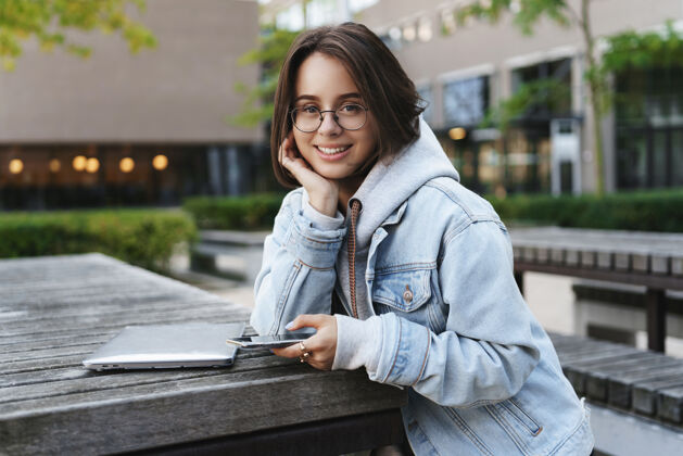 女孩近景写真：活泼可爱的年轻女学生 短发 倚在手掌上看着可爱的相机 面带微笑 坐在电脑旁 在户外使用笔记本电脑和手机笔记本电脑互联网准备好了