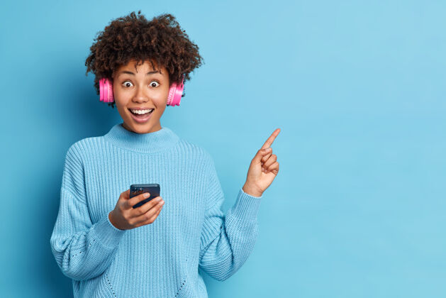 手指身着宽松针织毛衣的黑人女性手持现代手机 通过立体声耳机收听音乐耳机方向立体声