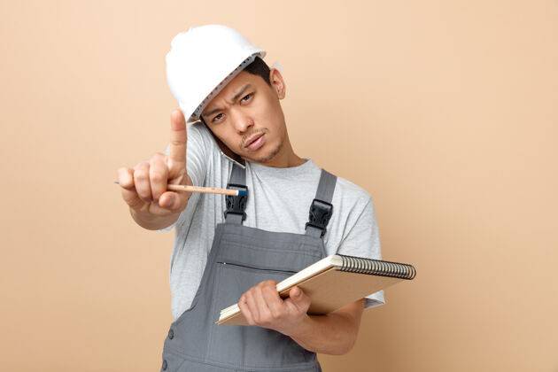 穿严格的年轻建筑工人戴着安全帽和制服拿着记事本和铅笔做握紧手势手势建筑严格