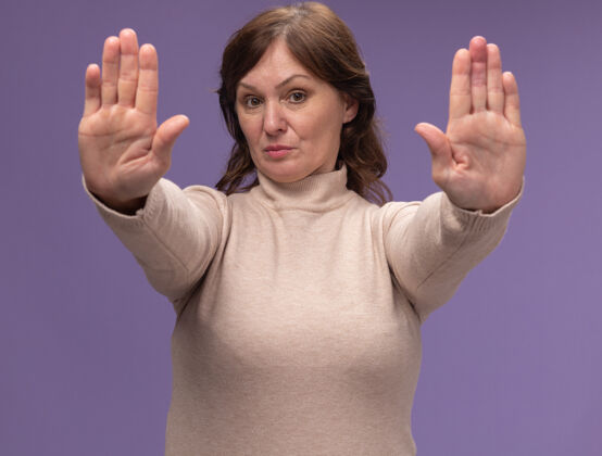 女人穿米色高领毛衣的中年妇女 严肃的脸 双手站在紫色的墙上做着停止的手势高领毛衣年龄中等