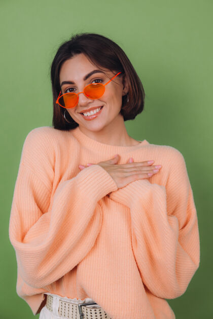 爱年轻时尚的女士穿着休闲桃色毛衣 戴着橘色眼镜 隔着绿橄榄色的墙正面双手合十放在胸前 心复制空间面部时尚温柔