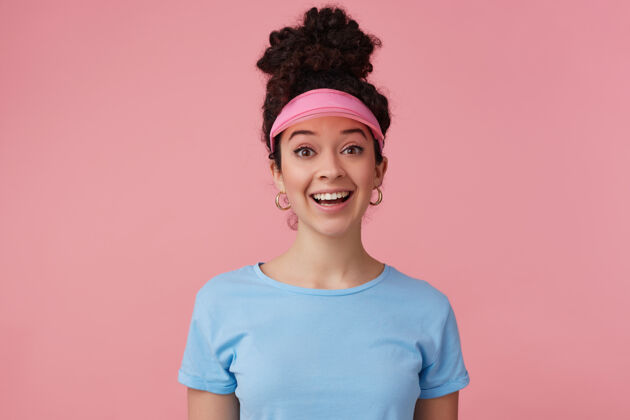 女性积极向上的女孩 快乐的女人 深色卷发发髻 戴着粉红色的帽舌 耳环和蓝色的t恤化妆情感概念卷曲漂亮情绪
