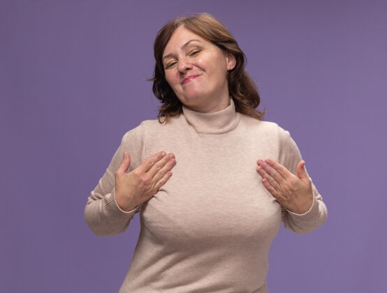 女人穿着米色套头衫的中年妇女站在紫色的墙上 高兴地指着自己高领毛衣中等年龄