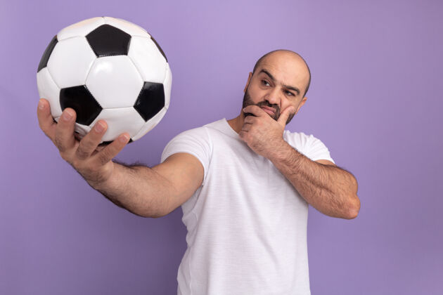 站一个留着胡子的男人 穿着白色t恤 手里拿着足球 脸上带着沉思的表情 站在紫色的墙上思考着表情球思考