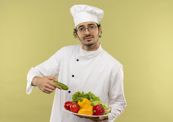 持有年轻的男厨师穿着厨师制服 戴着眼镜 盘子里放着蔬菜蔬菜烹饪盘子