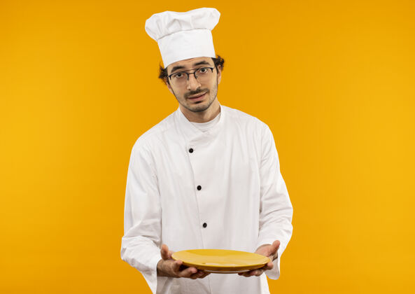 拿着高兴的年轻男厨师穿着厨师制服和眼镜托板年轻盘子厨师