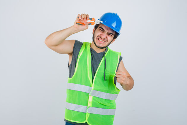 孩子年轻的建筑工人用钳子捏着头盔 穿着工作服展示握紧的拳头 看上去很有趣前视图时尚人青少年