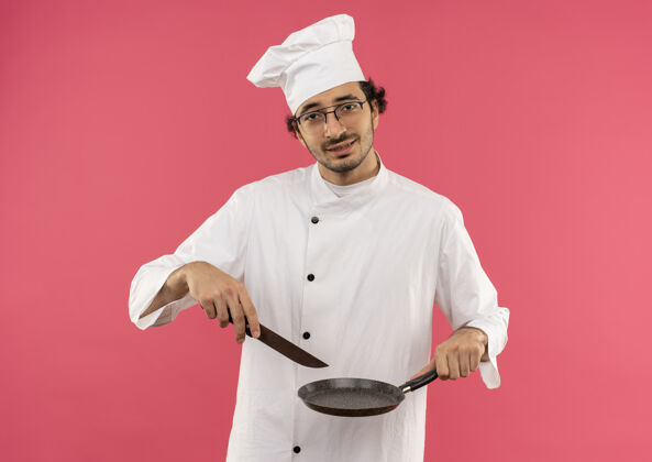 制服紧张的年轻男厨师穿着厨师制服 戴着眼镜 手里拿着煎锅和刀男年轻拿着