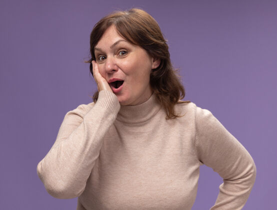 惊喜穿着米色高领套头衫的中年妇女站在紫色的墙壁上惊诧不已高领毛衣站女人