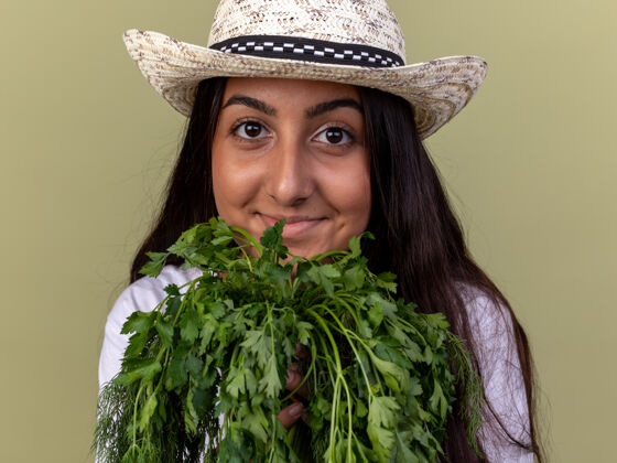 微笑戴着围裙和夏帽的快乐的年轻园丁女孩站在绿色的墙上 手里拿着新鲜的草药 脸上带着微笑新鲜草药站