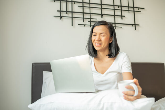 床快乐的休闲女人坐在家里的床上用笔记本电脑工作卧室女性舒适