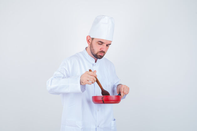 男性年轻的男厨师穿着白色制服 用木勺搅拌着饭菜 看上去很好奇 前视图美食木头烹饪