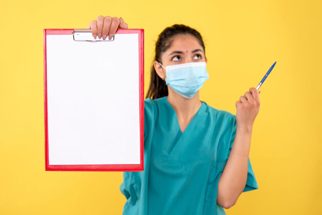 人物正面图：穿着制服的女医生拿着红色的剪贴板和笔站在黄色的背景上笔医院拿着
