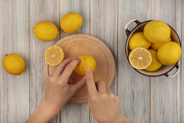 碗女性手在木制厨房板上切割柠檬的俯视图 刀子上放着柠檬 碗里的柠檬被隔离在灰色的木制表面上美味块健康