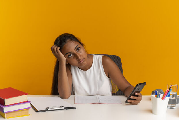 橙色年轻的女学生坐在书桌旁 把学习工具放在头上 手里拿着电话电话学校青少年