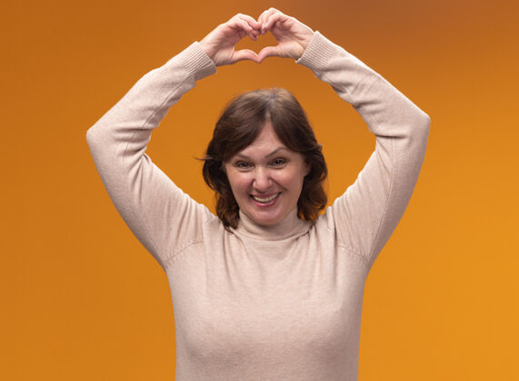年龄身穿米色套头衫的中年妇女站在橘色的墙上做着心形手势 脸上露出愉快的微笑高领毛衣制造姿势