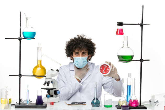 男性化学家正面图：男药剂师 穿着医疗服 戴着面具 在白色的空间里拿着红色的时钟实验室外套西装面具