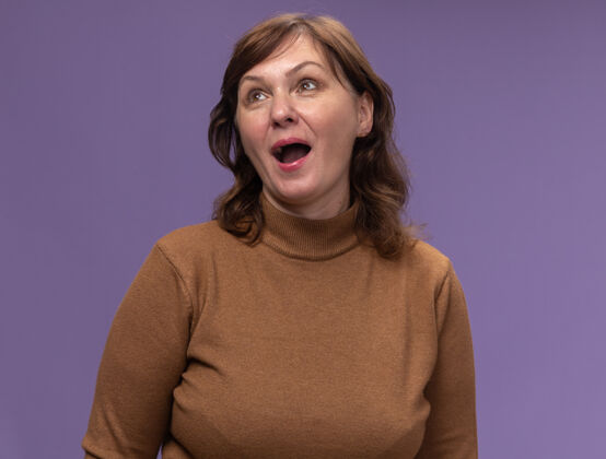 女人快乐的中年妇女穿着棕色高领毛衣 站在紫色的墙上 快乐地抬头仰望高领毛衣年龄中等