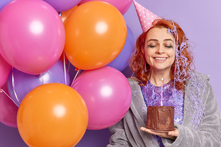 姿势高兴的女人有喜庆的心情捧着充气的彩色气球和生日蛋糕满意地闭上眼睛微笑愉快地表达真诚的情感高兴地接受生日的祝贺奶油女人蜡烛