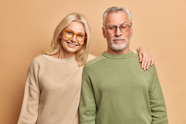 眼镜老年夫妇摆姿势为家庭画像拥抱微笑积极穿着眼镜套头衫站在棕色的工作室墙拥抱拥抱欢呼