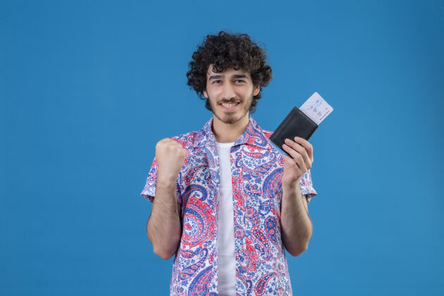 年轻面带微笑的年轻英俊的旅行家男子举着钱包和机票 高举拳头站在与世隔绝的蓝色墙壁上 留着复印空间钱包举起来机票