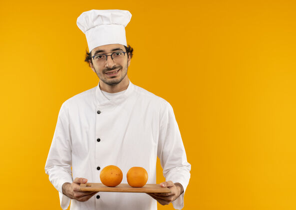 微笑面带微笑的年轻男厨师穿着厨师制服 戴着一副眼镜 手里拿着橘黄色的砧板 隔离在黄色的墙上男性烹饪厨师