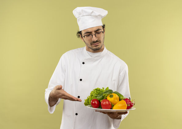 手想着年轻的男厨师穿着厨师制服 拿着眼镜 用手点着蔬菜放在盘上隔离在绿色的墙上拿着蔬菜年轻