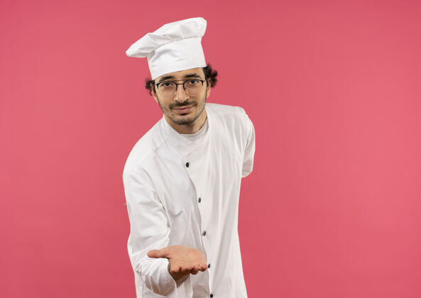 烹饪高兴的年轻男厨师穿着厨师制服 戴着眼镜 伸出手孤立在粉红色的墙上男性手持年轻