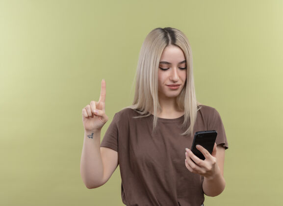 年轻年轻的金发女孩拿着手机看着它与孤立的绿色墙壁与复制空间举起手指电话手机举起