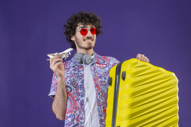 卷发微笑的年轻帅气的卷发旅行者戴着太阳镜拿着手提箱和模型飞机在隔离的紫色墙壁上复制空间手提箱男人穿