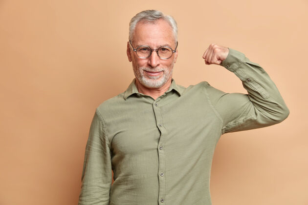 健身自信严肃的男人举起手臂展示肌肉自信他的力量穿着正式的衬衫对着棕色的墙壁摆姿势训练衬衫严肃