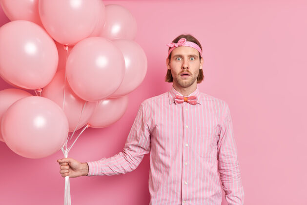 领结震惊惊讶的男人拿着一堆气球听到惊人的消息戴着头带优雅的衬衫打着蝴蝶结庆祝生日派对上的东西隔离在粉红色的墙上害怕周年纪念男