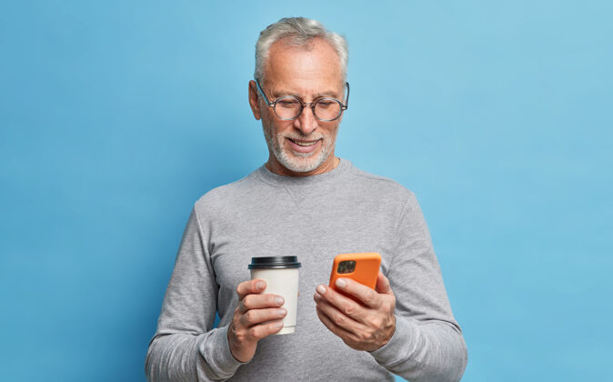 绅士开朗的现代老人用手机交流手机屏幕上的短信拿着纸杯咖啡卷轴穿着休闲服隔着蓝色的墙壁上网跳伞咖啡退休