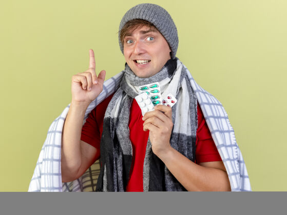 疾病令人印象深刻的年轻英俊的金发病男子戴着冬天的帽子和围巾包裹在格子布举行的药丸包指向橄榄绿的隔离墙围巾包格子