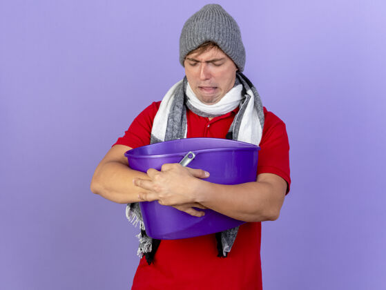 男人皱眉的年轻英俊的金发病男子戴着冬天的帽子和围巾拿着塑料桶闭着眼睛有恶心隔离在紫色墙上的复制空间穿水桶英俊