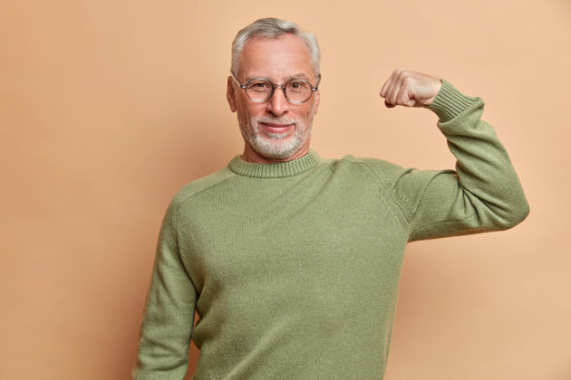 年龄自信满满的白发男子举起手臂 展示肌肉 在健身房定期训练后展示效果戴着眼镜和跳投 隔着棕色的墙壁 为自己感到骄傲自信强壮力量