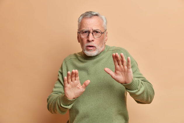 困惑摄影棚拍摄的一个害怕的白发男人保持着手掌的防御姿态 要求不要靠近 看到恐惧症穿着休闲毛衣 戴着眼镜隔着棕色的墙男性成人眼镜