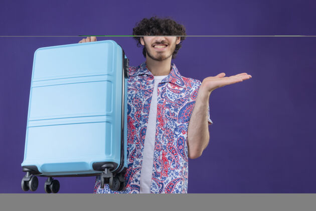 年轻微笑的年轻英俊的卷发旅行者男子拿着手提箱 在孤零零的紫色墙上空手展示帅气男人卷发