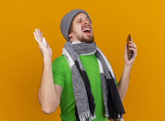 穿愤怒的年轻英俊的斯拉夫病夫戴着冬天的帽子和围巾拿着手机和餐巾闭着眼睛孤立在橙色的墙上餐巾愤怒男人