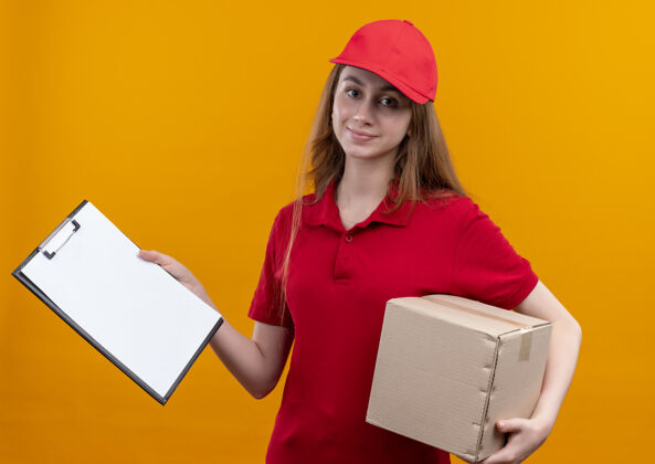 年轻穿着红色制服的年轻送货女孩把盒子和剪贴板放在孤立的橙色墙上拿着剪贴板女人