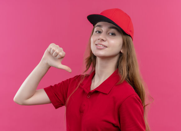 指着身着红色制服 面带微笑的年轻送货员在孤立的粉色墙上指着自己交货年轻女人