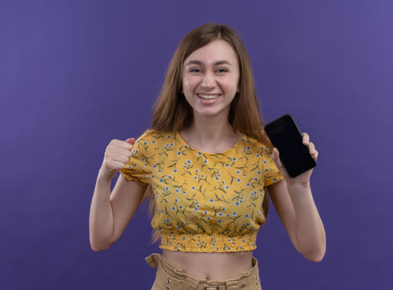 女孩微笑的年轻女孩拿着手机 举起拳头对着孤零零的紫色墙壁握着微笑电话