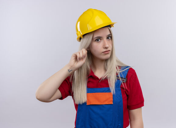 手指穿着制服的年轻金发建筑工女孩把手指放在耳朵上看着孤立的白色墙壁工程师建筑工人金发