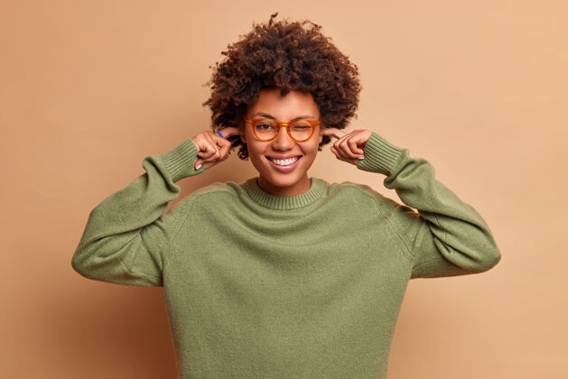 肖像积极的卷发女人开心地笑着塞住耳朵避免很大声的音乐戴着透明的眼镜和毛衣有快乐的心情隔着棕色的墙套头衫休闲高兴