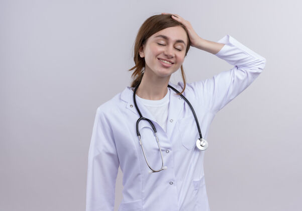 女人面带微笑的年轻女医生 穿着医用长袍 戴着听诊器 将手放在头上 放在隔离的白色墙壁上 留有复印空间年轻医生穿