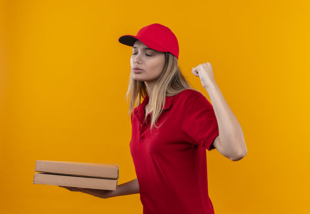 关闭年轻的送货女孩闭着眼睛 穿着红色制服 拿着披萨盒 在橙色的墙上显示“是”的手势披萨手势帽子