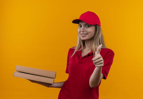 披萨微笑着的年轻送货女孩穿着红色制服 戴着帽子 手里拿着披萨盒 她的大拇指孤立地竖在橙色的墙上微笑拿着送货
