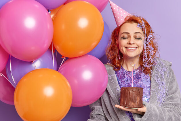 姿势高兴的女人有喜庆的心情捧着充气的彩色气球和生日蛋糕满意地闭上眼睛微笑愉快地表达真诚的情感高兴地接受生日的祝贺奶油女人蜡烛