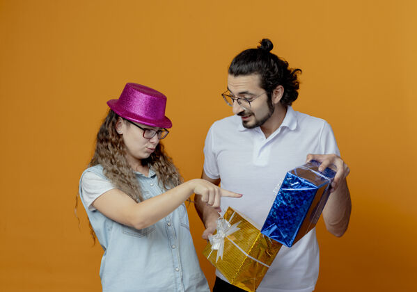 年轻人一对年轻夫妇戴着粉红色帽子的女孩指着男孩手中的礼物孤立在橙色的墙上女孩帽子点