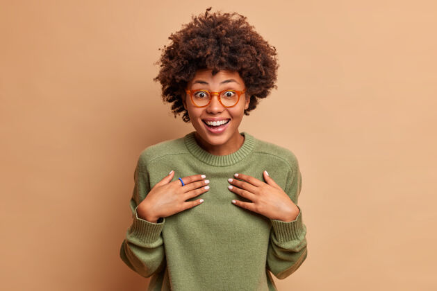 非洲快乐女人的横拍手放在胸前 微笑着 收到意想不到的礼物时反应愉快戴着透明眼镜和套头衫隔着棕色的墙可爱眼镜休闲